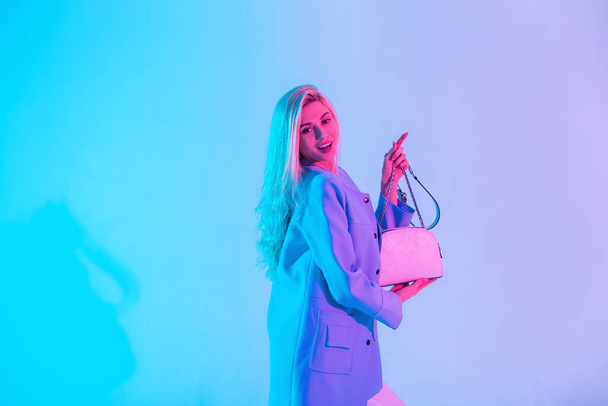 Moda mavisi ceketli, neşeli, çekici ve çekici sarışın bir kadın, neon ışıklı arka planda bir stüdyoda şık bir el çantası tutuyor. - Fotoğraf, Görsel