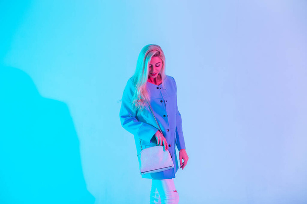Ευτυχισμένη νεαρή ξανθιά κοπέλα σε ένα μοντέρνο μπλε κομψό σακάκι με ένα λευκό κομψό δερμάτινη τσάντα στέκεται στο στούντιο για νέον φως με ροζ - Φωτογραφία, εικόνα