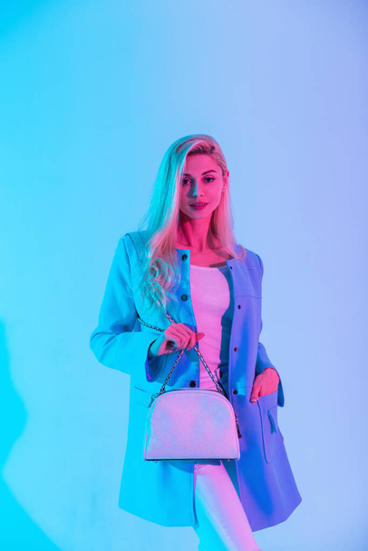 Mujer de negocios hermosa de moda en ropa elegante brillante y chaqueta con un elegante bolso blanco en un estudio con luz pastel creativa - Foto, imagen