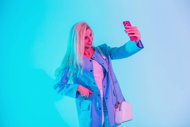 Moda sektörü kıyafetleri içinde sevimli bir gülüşü olan mutlu sarışın kadın çantayla görünüyor ve spor ceketi stüdyodaki parlak telefonunda çok renkli bir ışıkla selfie çekiyor. - Fotoğraf, Görsel