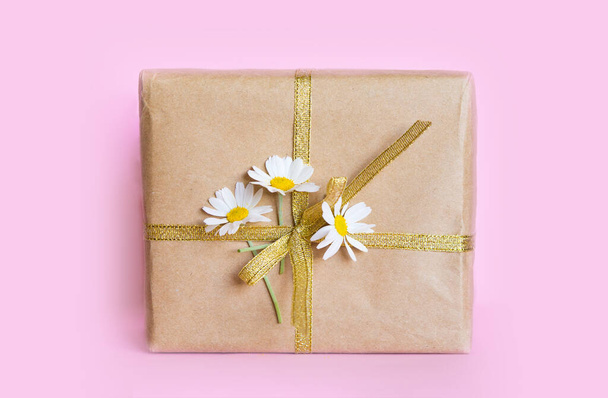 Cadeau dans une boîte attachée avec un ruban d'or et un bouquet de marguerites sur un fond rose pastel. Cadeau pour le 8 mars ou la fête des mères. - Photo, image