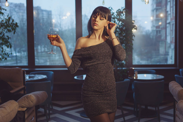 Beautiful woman with a glass of wine stands near the evening window. The woman's shoulders are open and she looks sexy. - Фото, зображення