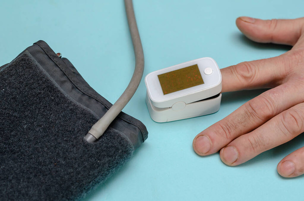 Egy kéz fehér oximéterrel a mutatóujján. A közelben van egy digitális vérnyomásmérő. Egy érett férfi méri a kapilláris vér oxigénszintjét és a pulzusát. Szelektív fókusz. - Fotó, kép
