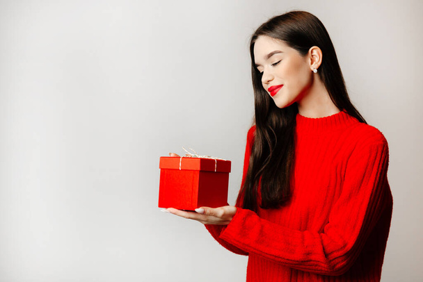 Attractive girl with black hair and red lips. The girl is staying against the light background with a present in her hands - Photo, Image