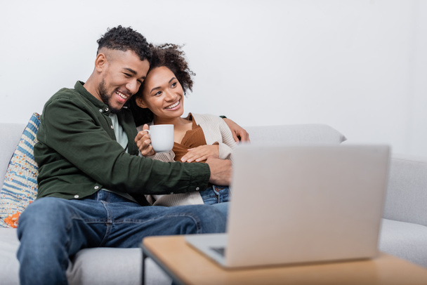 cheerful african american man hugging pregnant wife near blurred laptop - Fotó, kép