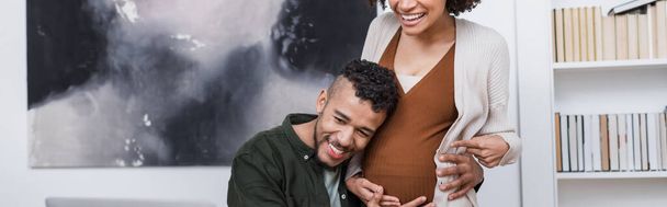 vrolijk zwanger Afrikaans amerikaanse vrouw wijzend op buik in de buurt gelukkig echtgenoot, banner - Foto, afbeelding