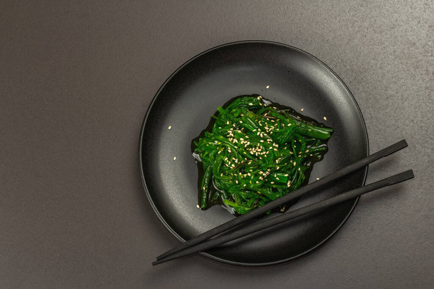 Wakame seaweed salad with sesame seeds and chili pepper in a bowl. Trendy hard light, dark shadow. Black stone concrete background, flat lay, top view - Foto, Bild