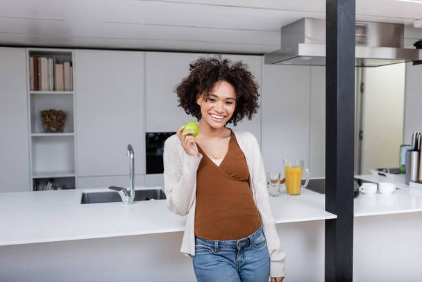 Χαρούμενη έγκυος Αφροαμερικανή γυναίκα κρατώντας πράσινο μήλο στην κουζίνα  - Φωτογραφία, εικόνα