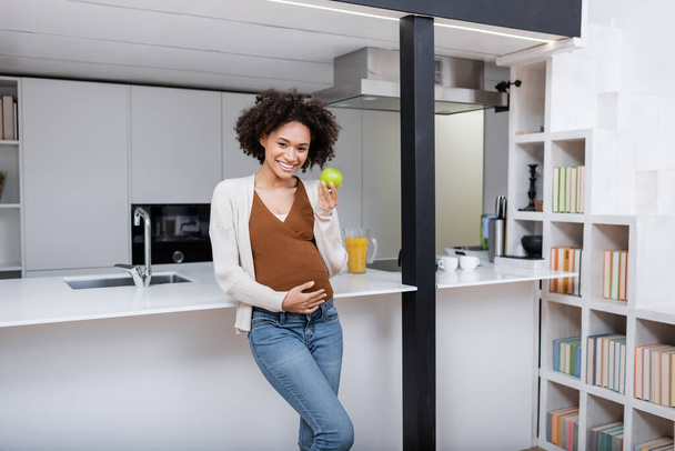 happy pregnant african american woman holding green apple in kitchen  - Foto, Bild