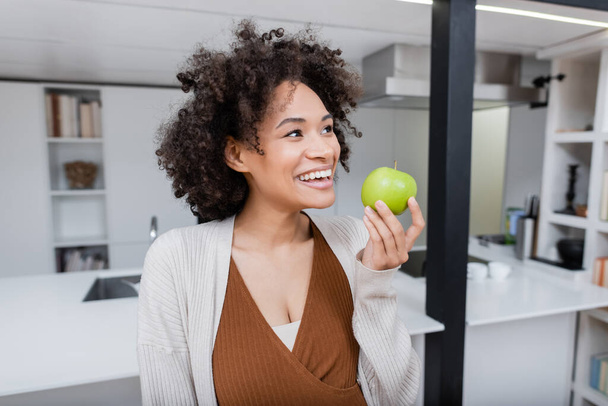 wesoła ciężarna Afroamerykanka trzyma dojrzałe jabłko w kuchni  - Zdjęcie, obraz