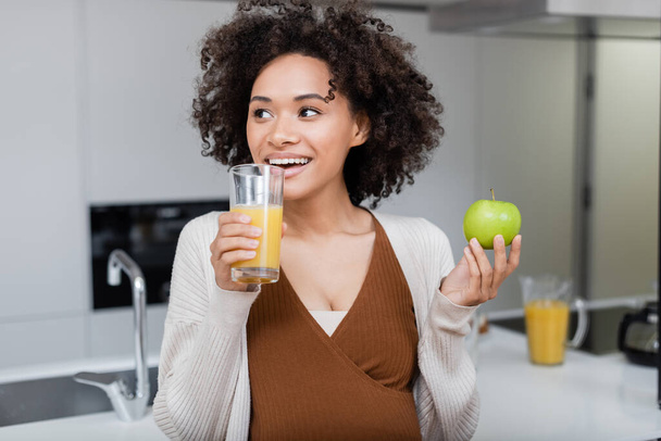 alegre embarazada africana americana mujer sosteniendo verde manzana y vaso de jugo de naranja en cocina  - Foto, Imagen