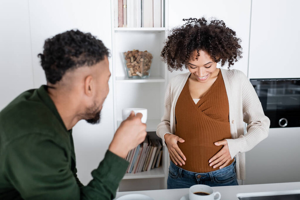 desenfocado africano americano hombre mirando embarazada esposa mientras celebración taza con café en cocina  - Foto, imagen