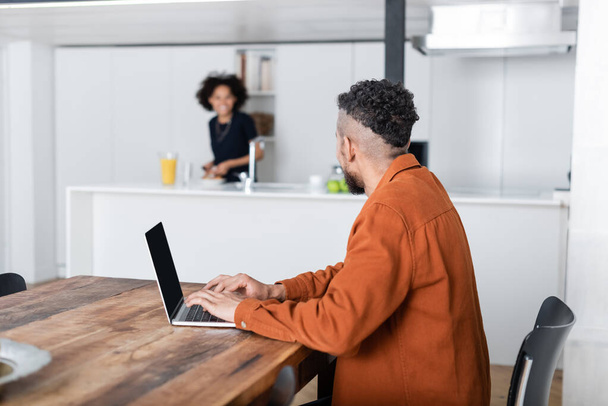 african american man using laptop while working from home and looking at blurred girlfriend in kitchen  - Foto, Imagen