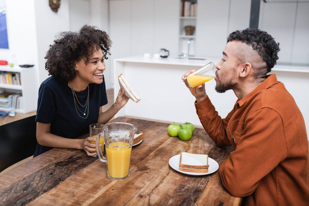 happy african american woman holding sandwich while boyfriend drinking orange juice  - Fotografie, Obrázek