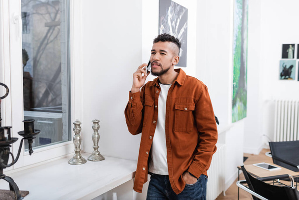 lockiger afrikanisch-amerikanischer Mann telefoniert, während er mit der Hand in der Tasche in einer modernen Wohnung steht  - Foto, Bild