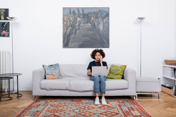 curly african american woman sitting on couch and talking on smartphone near laptop  - Photo, image