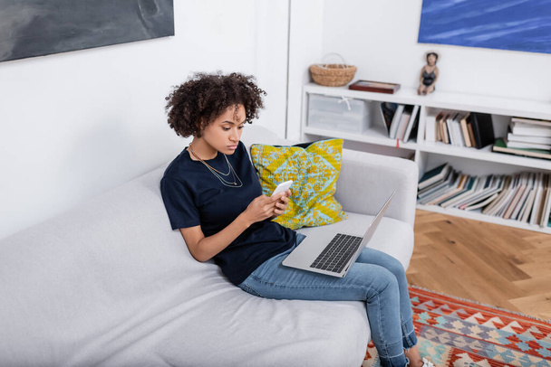 curly african american woman using cellphone near laptop while sitting on sofa - Fotoğraf, Görsel