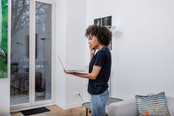 curly african american woman talking on smartphone and holding laptop in living room - Foto, Bild