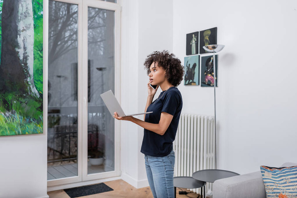 young african american woman talking on smartphone and holding laptop in living room - Photo, Image