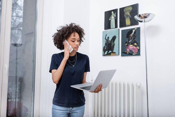 Genç Afro-Amerikalı kadın cep telefonuyla konuşuyor ve modern apartmanda dizüstü bilgisayar tutuyor.  - Fotoğraf, Görsel