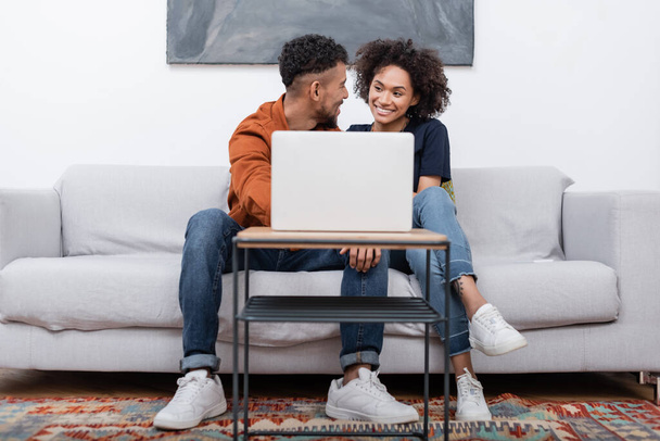 happy young african american couple smiling while looking at each other near laptop in modern apartment   - Φωτογραφία, εικόνα