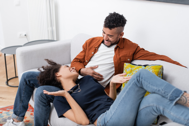 pleased african american couple looking at each other while resting on couch in modern apartment  - Photo, image