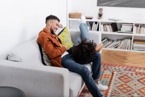 Χαρούμενος νεαρός Αφροαμερικάνος που κοιτάζει την κοπέλα του να αναπαύεται στον καναπέ στο μοντέρνο διαμέρισμα  - Φωτογραφία, εικόνα