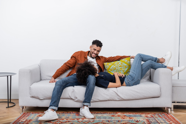 Χαρούμενος Αφροαμερικάνος που κοιτάζει την κοπέλα του που αναπαύεται στον καναπέ στο μοντέρνο διαμέρισμα  - Φωτογραφία, εικόνα