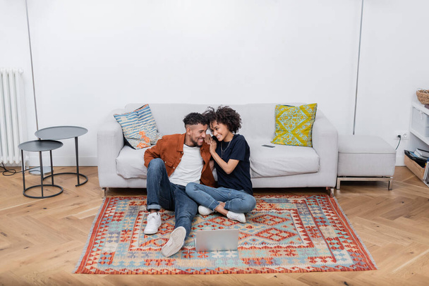 joyful african american couple sitting on carpet and looking at each other near laptop and smartphone on sofa - Foto, immagini