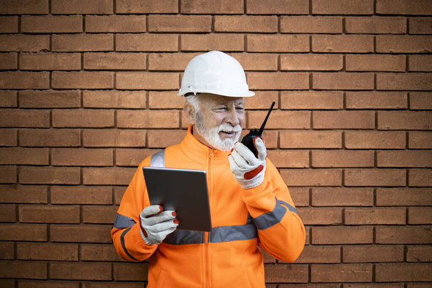 Portrait of construction worker supervisor in safety equipment and hard had standing against brick wall, holding tablet and communicating via radio at construction site. - Foto, immagini