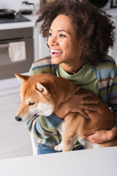 mujer afroamericana llena de alegría mirando hacia otro lado mientras abraza al perro shiba inu en la cocina - Foto, imagen