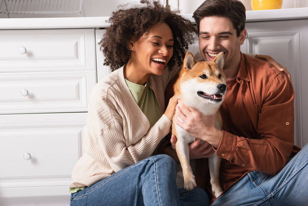 jovem e feliz casal interracial se divertindo com shiba inu cão na cozinha - Foto, Imagem