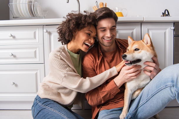pleased interracial couple hugging shiba inu dog while sitting on kitchen floor - Photo, Image