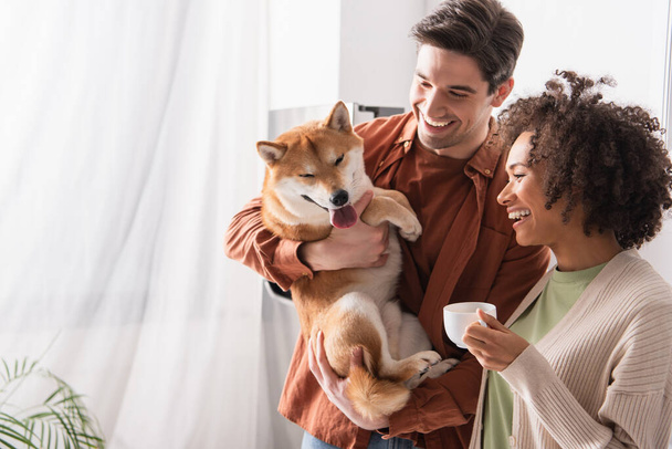 アフリカ系アメリカ人女性の近くで面白い柴犬を持っている陽気な男コーヒーを飲みながら - 写真・画像