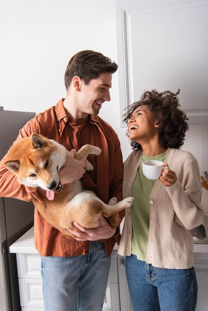 funny shiba inu dog sticking out tongue near happy interracial couple smiling at each other in kitchen - Foto, Bild