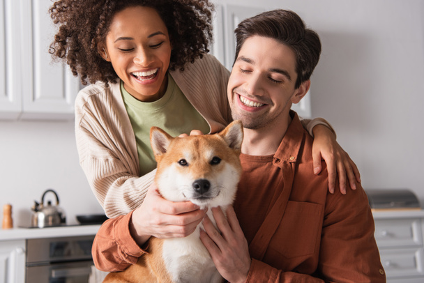 shiba inu dog looking at camera near pleased multiethnic couple in kitchen - Photo, Image