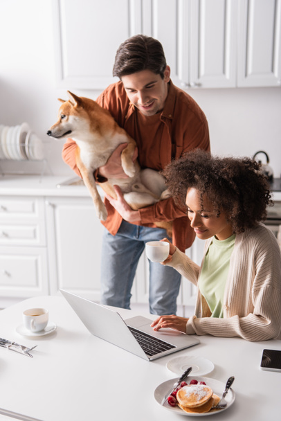 young man with shiba inu dog looking at laptop near smiling african american girlfriend - Fotó, kép