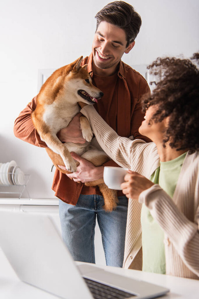 african american woman with coffee cup stroking shiba inu dog in hands of smiling boyfriend - Фото, изображение