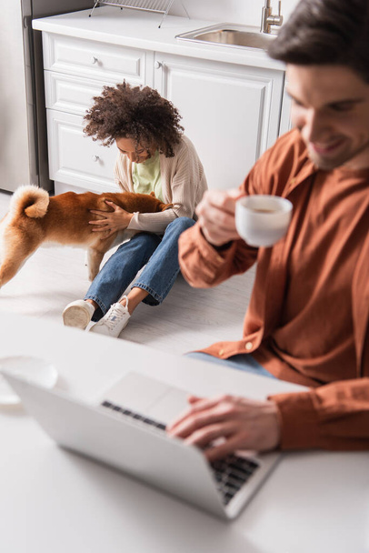 African american woman playing with shiba inu near blurred boyfriend using laptop and holding cup in kitchen  - Φωτογραφία, εικόνα
