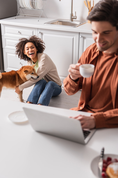 Positive african american woman playing with shiba inu near blurred boyfriend with cup using laptop in kitchen  - 写真・画像