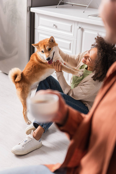 Smiling african american woman playing with shiba inu near blurred man with cup in kitchen  - Foto, Imagem