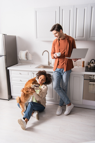 Smiling man holding cup and laptop near african american girlfriend playing with shiba inu in kitchen  - Photo, Image