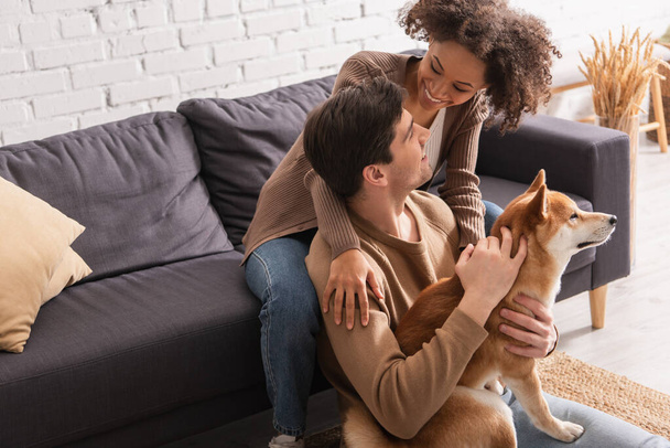 Happy interracial couple petting shiba inu and looking at each other at home  - Photo, Image