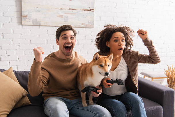 KYIV, UKRAINE - DECEMBER 22, 2021: Excited interracial couple showing yes gesture while playing video game near shiba inu at home  - Photo, image