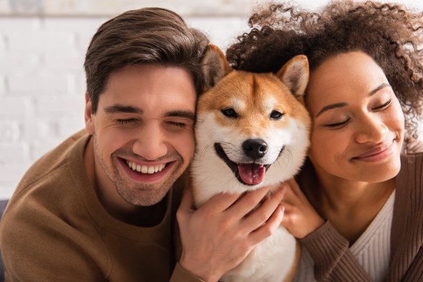 Cheerful man with closed eyes petting shiba inu near african american girlfriend at home  - Foto, Imagen