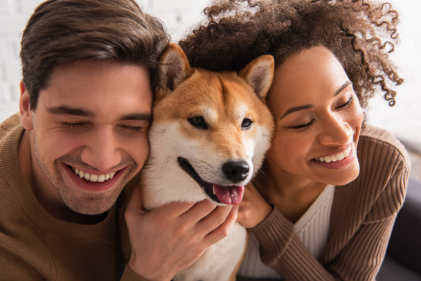 Smiling multiethnic couple with closed eyes petting shiba inu at home  - Photo, Image
