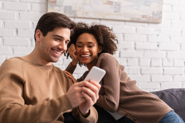 Man using smartphone near smiling african american girlfriend on couch at home  - Φωτογραφία, εικόνα