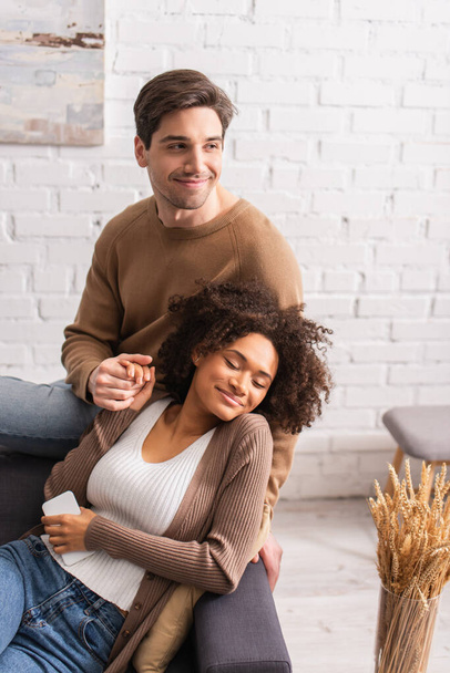 Smiling man holding hand of african american girlfriend with cellphone on couch  - Photo, Image