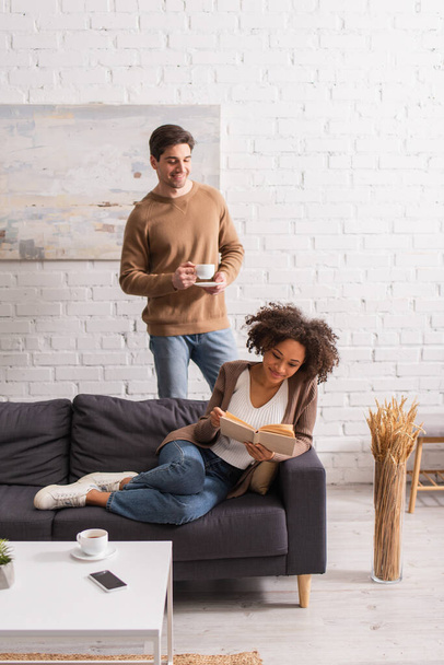 Man holding coffee cup near african american girlfriend reading book on couch at home  - Foto, afbeelding