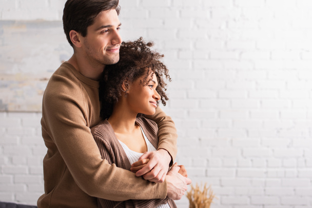 Smiling man hugging curly african american girlfriend at home  - Photo, Image